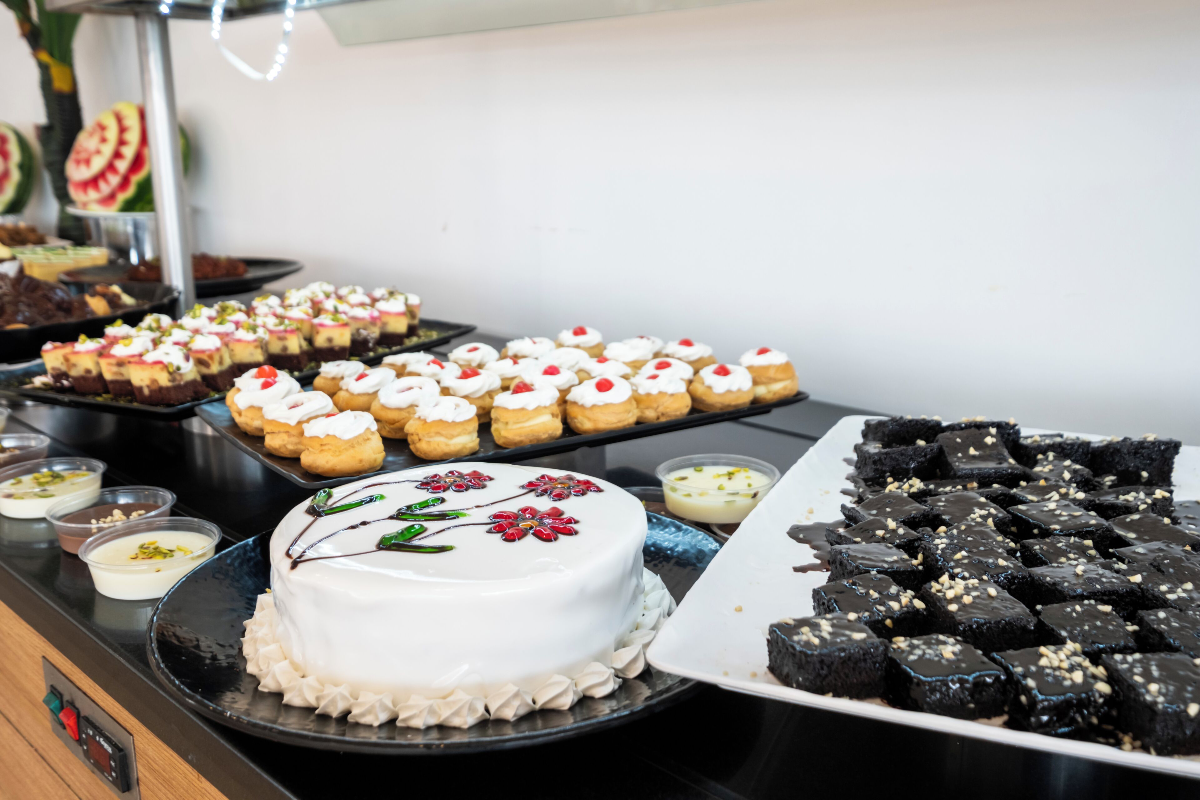 En buffé med olika sorters desserter, inklusive en vit tårta med blomsterdekorationer, muffins med rött strössel och chokladöverdragna godsaker.