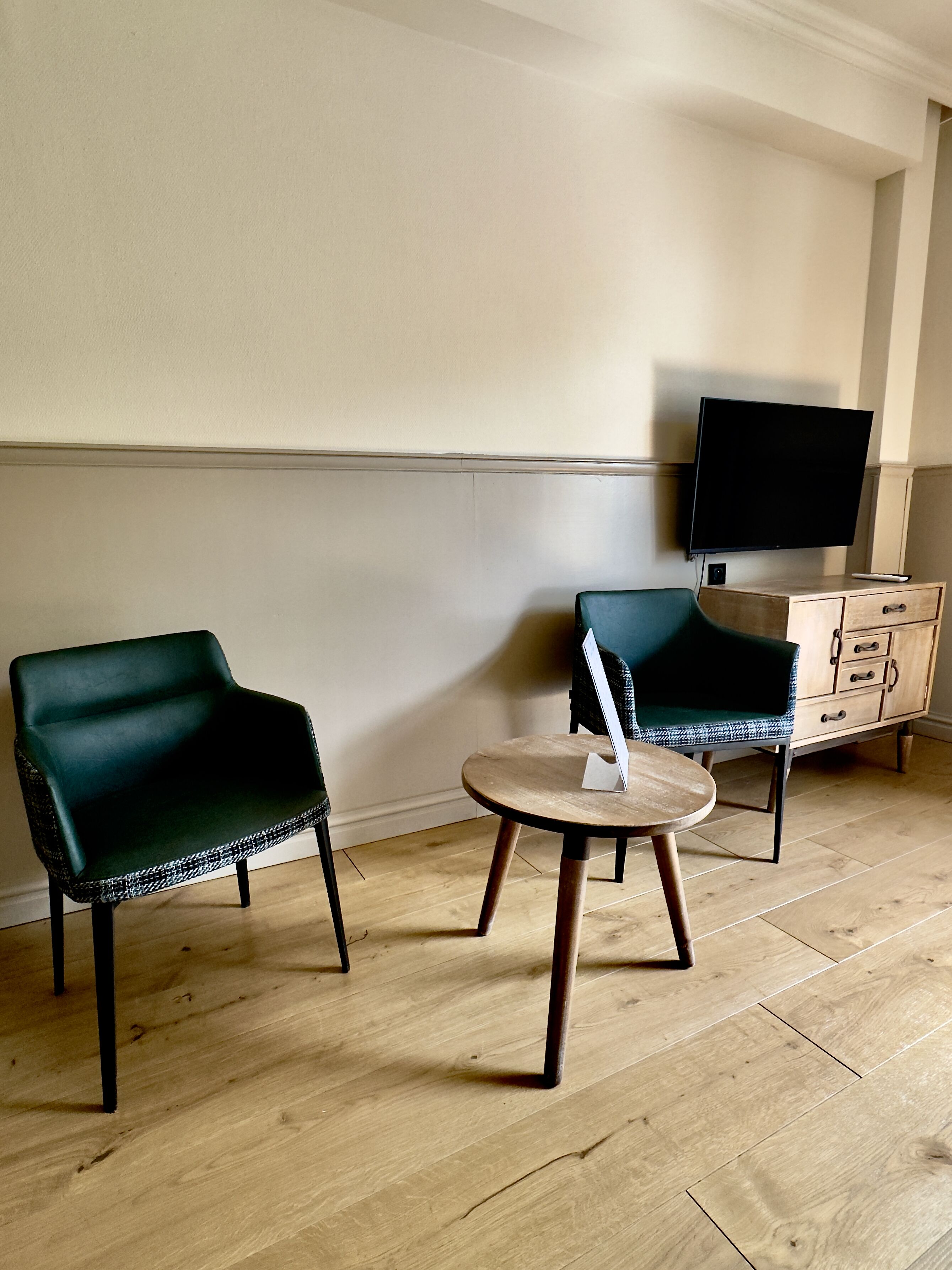 Een kamer met twee donkergroene fauteuils, een klein rond tafeltje en een houten kast met lades. Een flatscreen televisie hangt aan de muur.