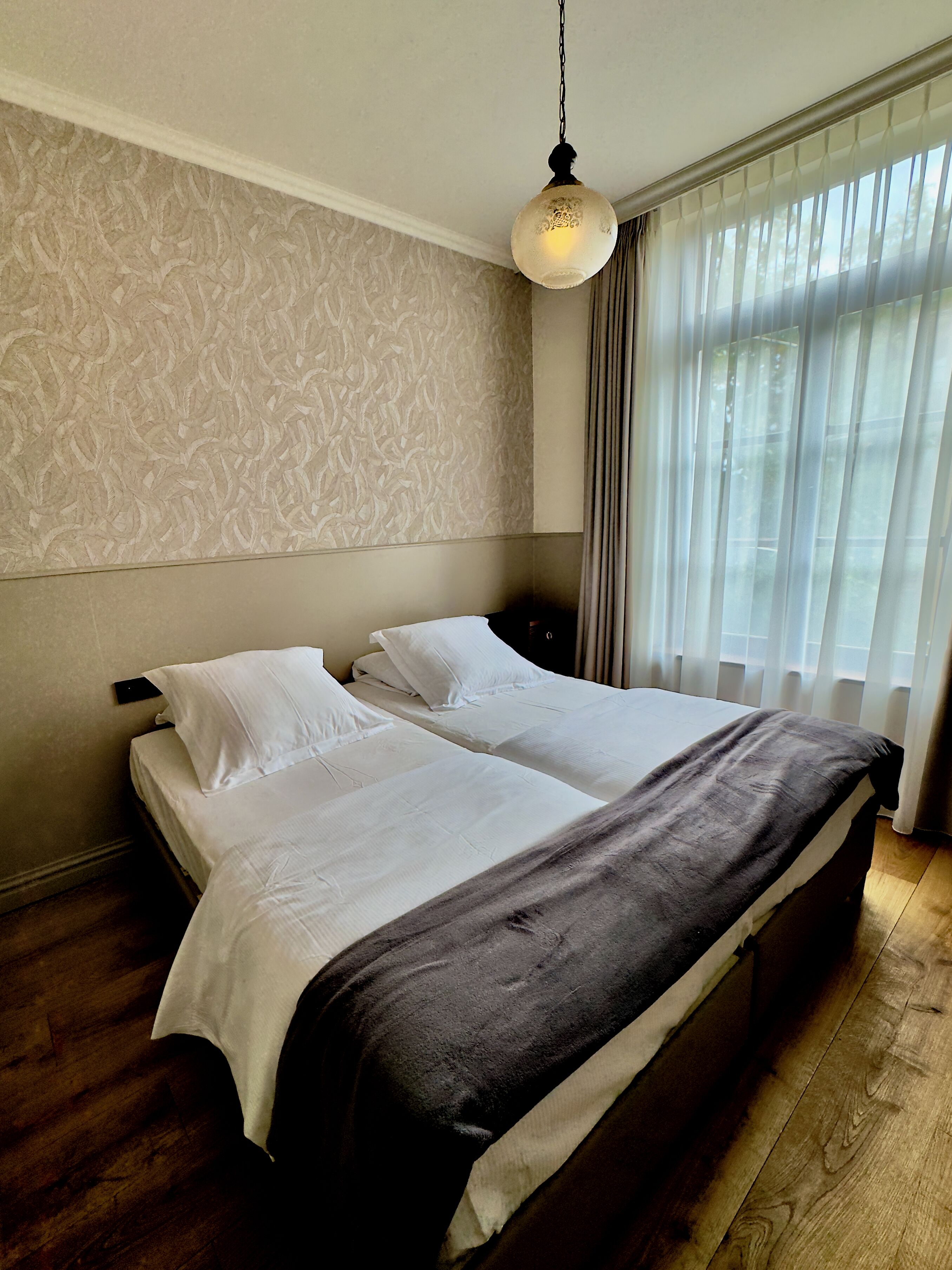 A double bed with white linens, a dark throw blanket, and a hanging light fixture.