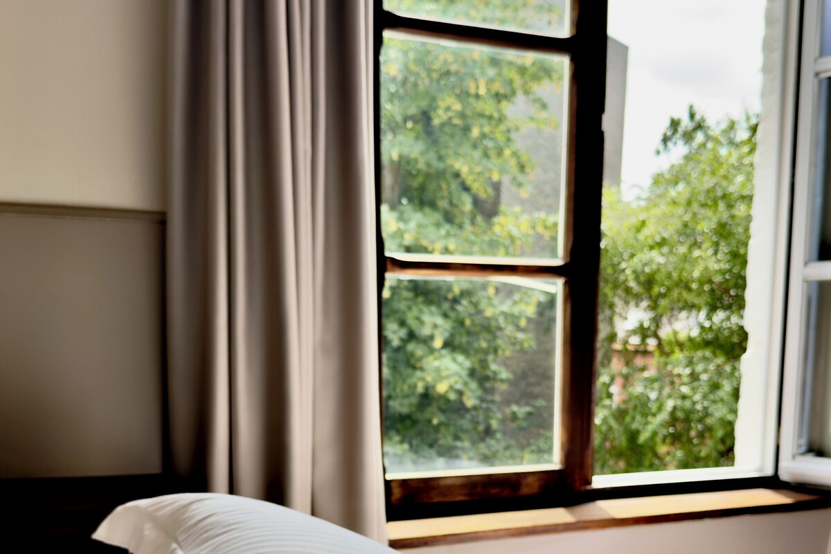 A bedroom with a bed, a window with curtains, and a view of greenery outside.