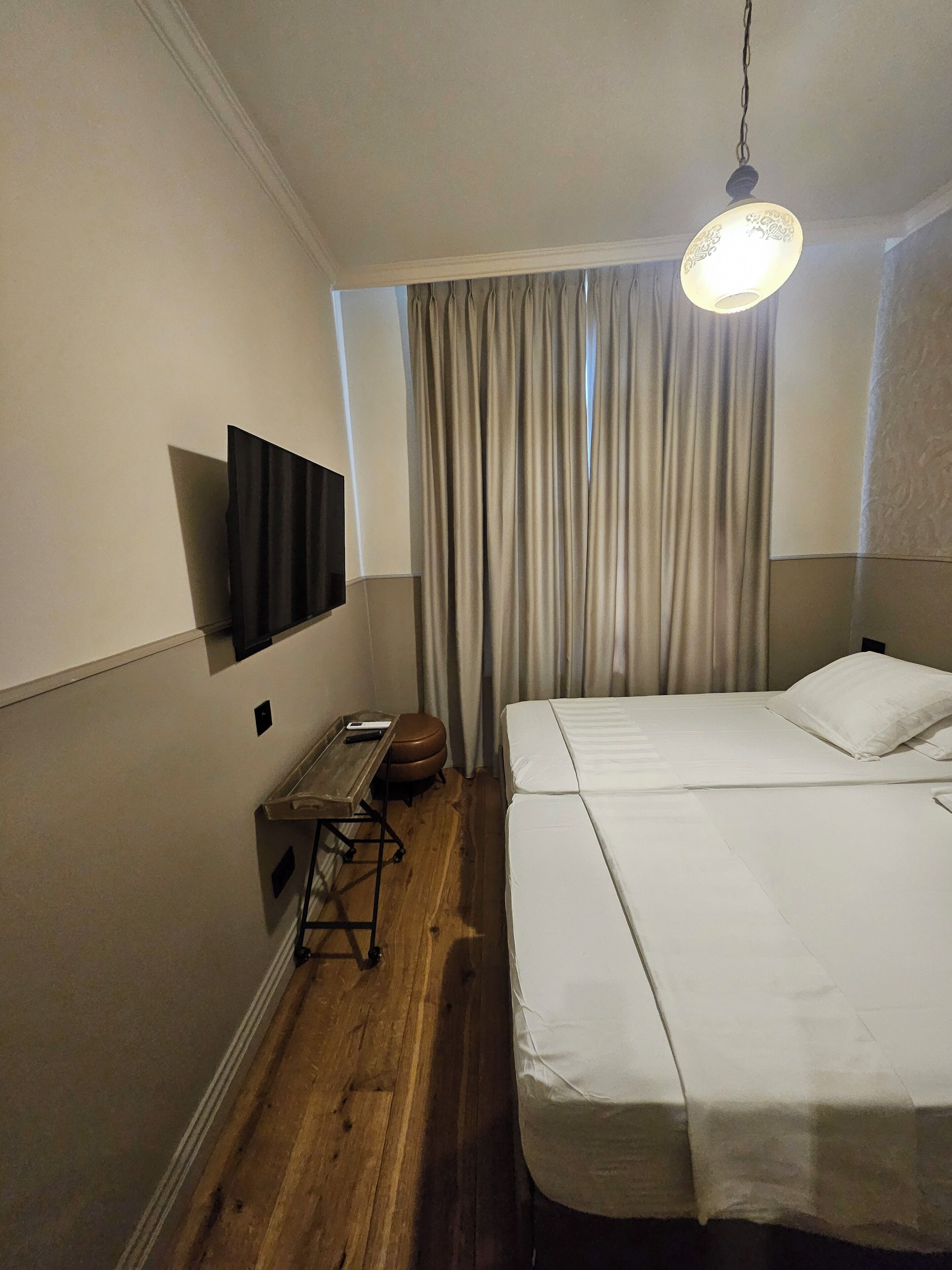 Quarto de hotel com cama, televisão de tela plana na parede, piso de madeira e janela com cortinas.
