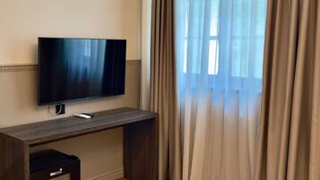Ein Zimmer mit einem an einer Holztischplatte montierten Flachbildfernseher, einem kleinen schwarzen Gerät auf der Tischplatte und einem Fenster mit durchsichtigen Vorhängen.