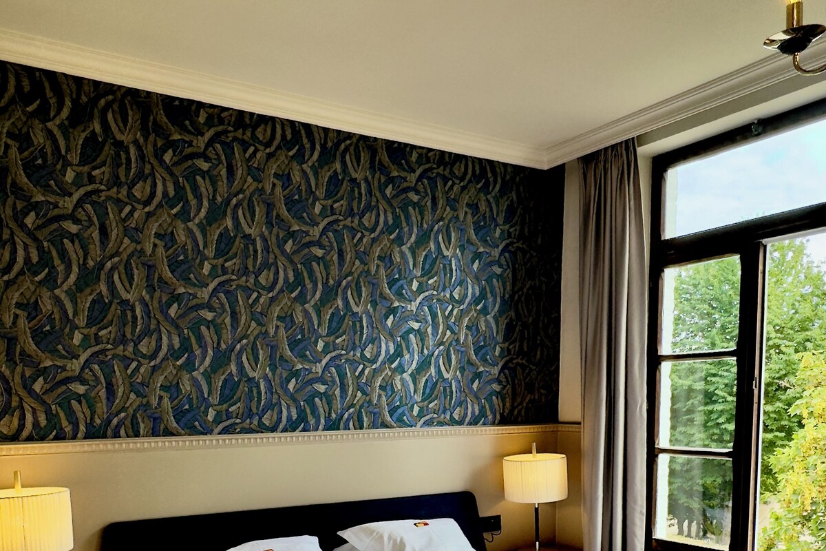A hotel room with a large bed, two bedside lamps, a patterned wall, and a window with a view of greenery.