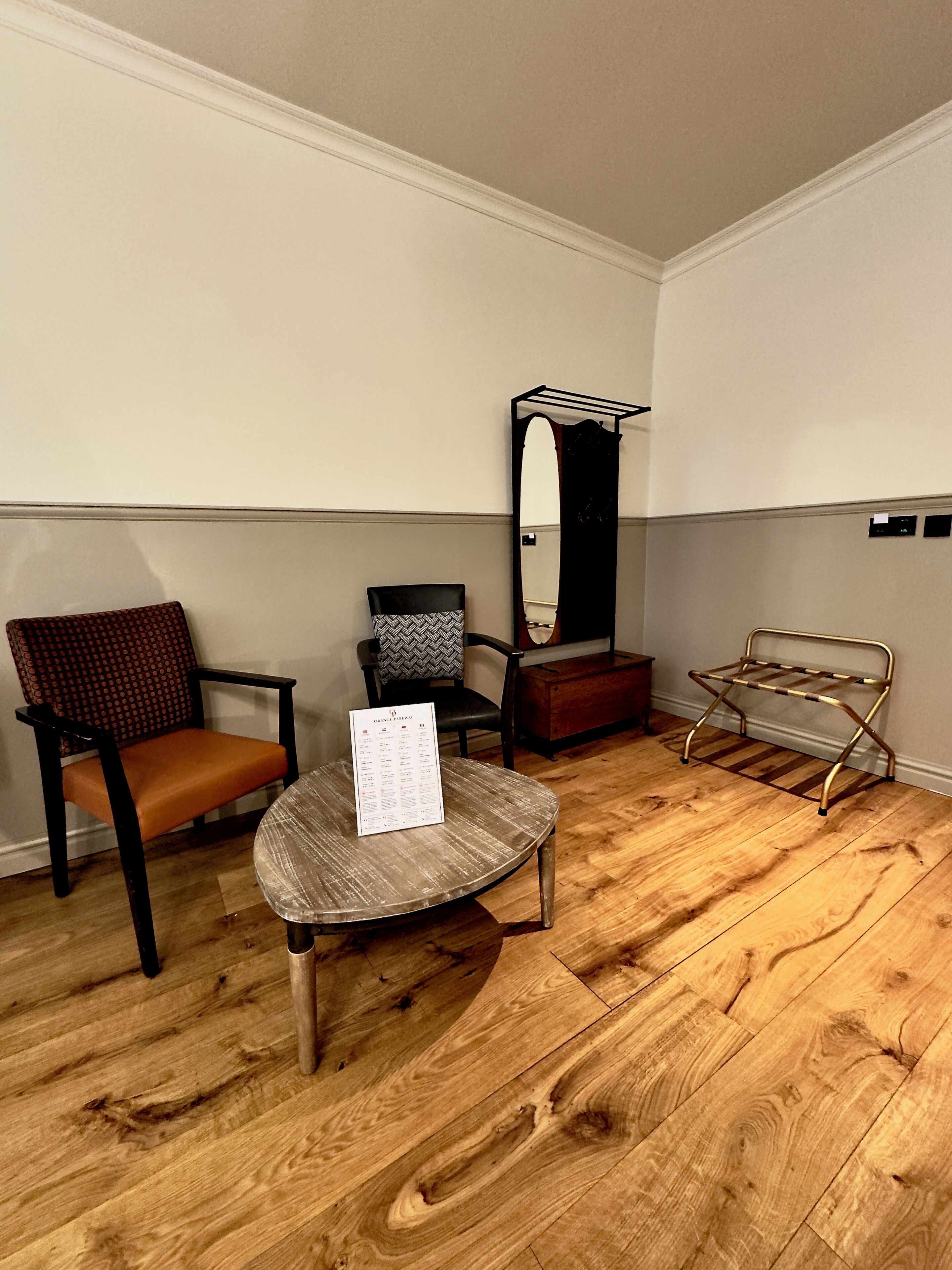 A room with a wooden floor, a round table with a menu, a black chair, a patterned chair, a mirror, and a metal rack.