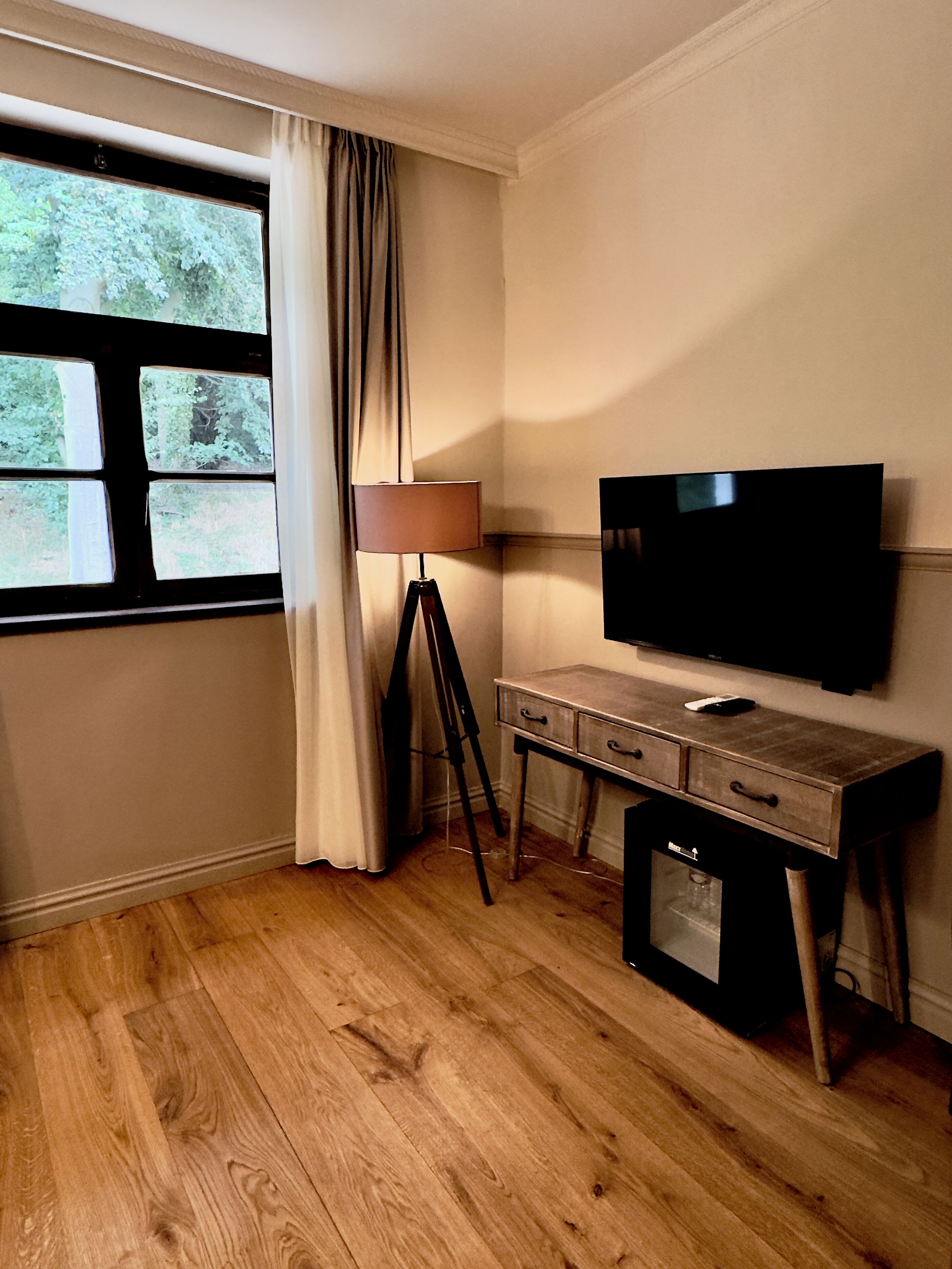 A room with a large window, a desk with drawers, a lamp, and a flat-screen TV mounted on the wall.