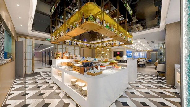 A modern hotel lobby with a central bar area, geometric patterned flooring, and a high ceiling with exposed structural elements. - Banana Design Residence Guangzhou Haizhu Changgang Metro Branch (Guangzhou)