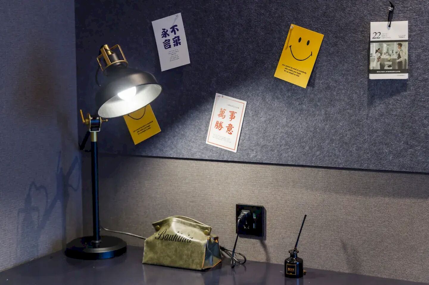 Et skrivebord med en skrivebordslampe, en radio samt en væg med notater og en kalender.