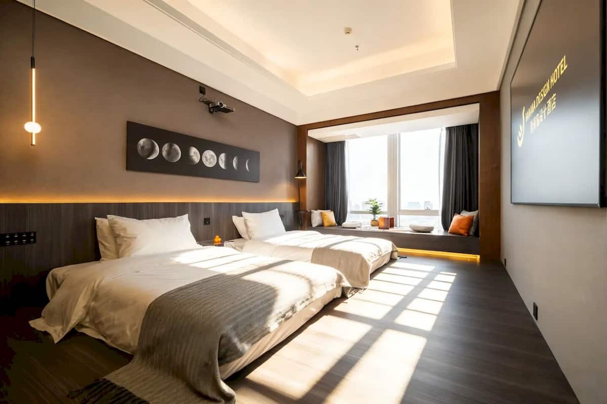 Hotellihuone, jossa on kaksi sänkyä, seinälle kiinnitetty taideteos, jossa on kuvattuna kuita, suuri ikkuna verhoineen sekä seinälle kiinnitetty kyltti.