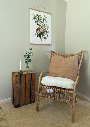 A wicker chair with a white cushion, a small wooden side table with a candle and a plant, and a framed botanical illustration on the wall. - Unique accommodations close to nature in northern Sweden (Gran)