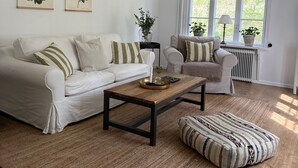 Un salon avec un canapé blanc, une table basse en bois et un pouf à motifs. Deux reproductions botaniques encadrées ornent le mur, une fenêtre offre une vue sur la verdure et un radiateur est installé.