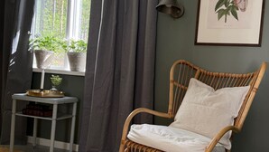 Une chaise en osier avec un coussin, une petite table avec un plateau et une bougie, et une estampe botanique encadrée accrochée au mur.