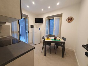 A compact kitchen with a refrigerator, a dining table with chairs, and a flat-screen TV mounted on the wall. - Oasis of Peace: three-room apartment with private garden a few steps from the sea (Fregene)
