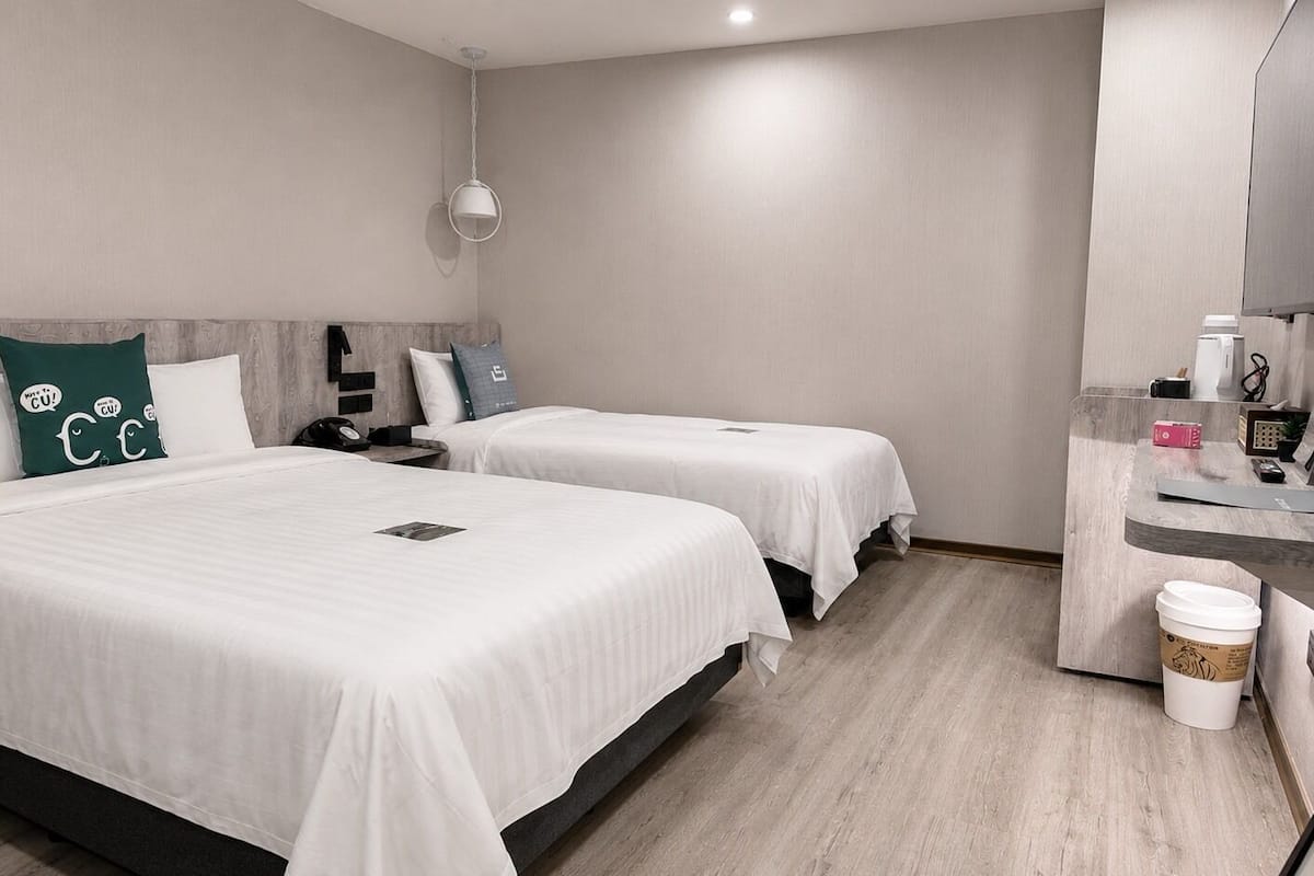 A hotel room with two beds, a desk, a trash bin, and a hanging light fixture.