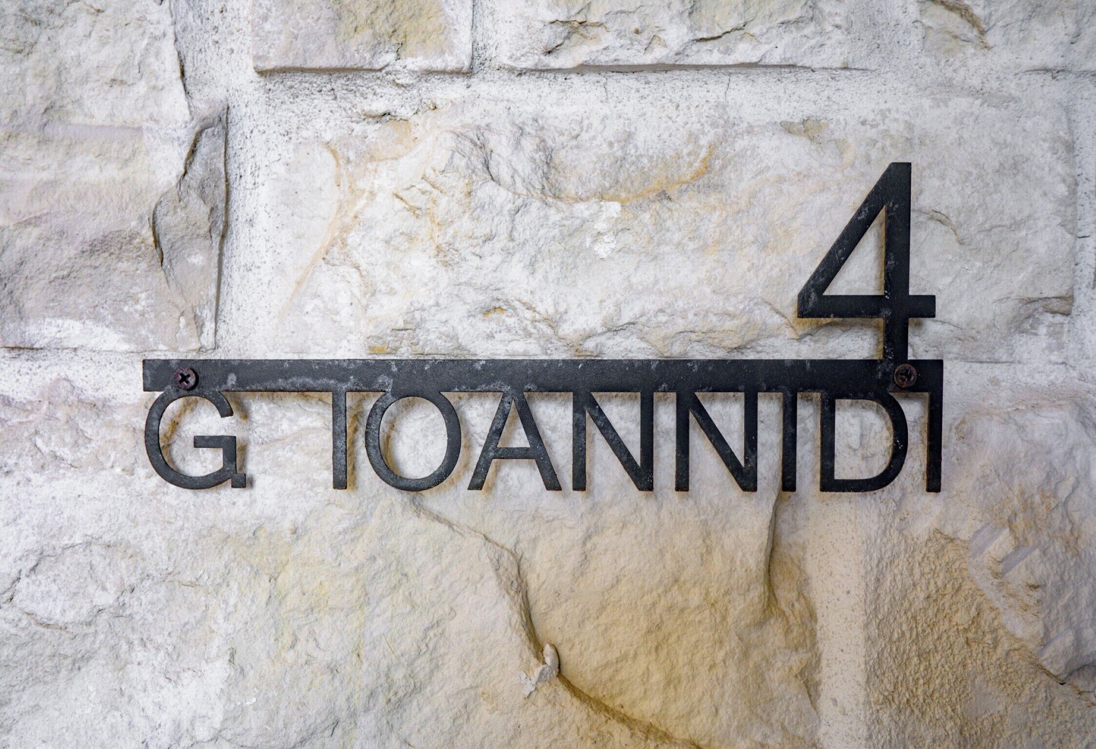 Un cartello metallico con la scritta 'G TOANNIDI' e il numero '4' su un muro di pietra.