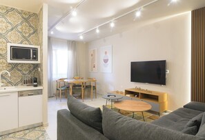 A modern living room with a sofa, a TV, a small table, and a kitchen area with a microwave. - A modern apartment in the center of discreet luxury and comfort 30m from the sea (Ierapetra)