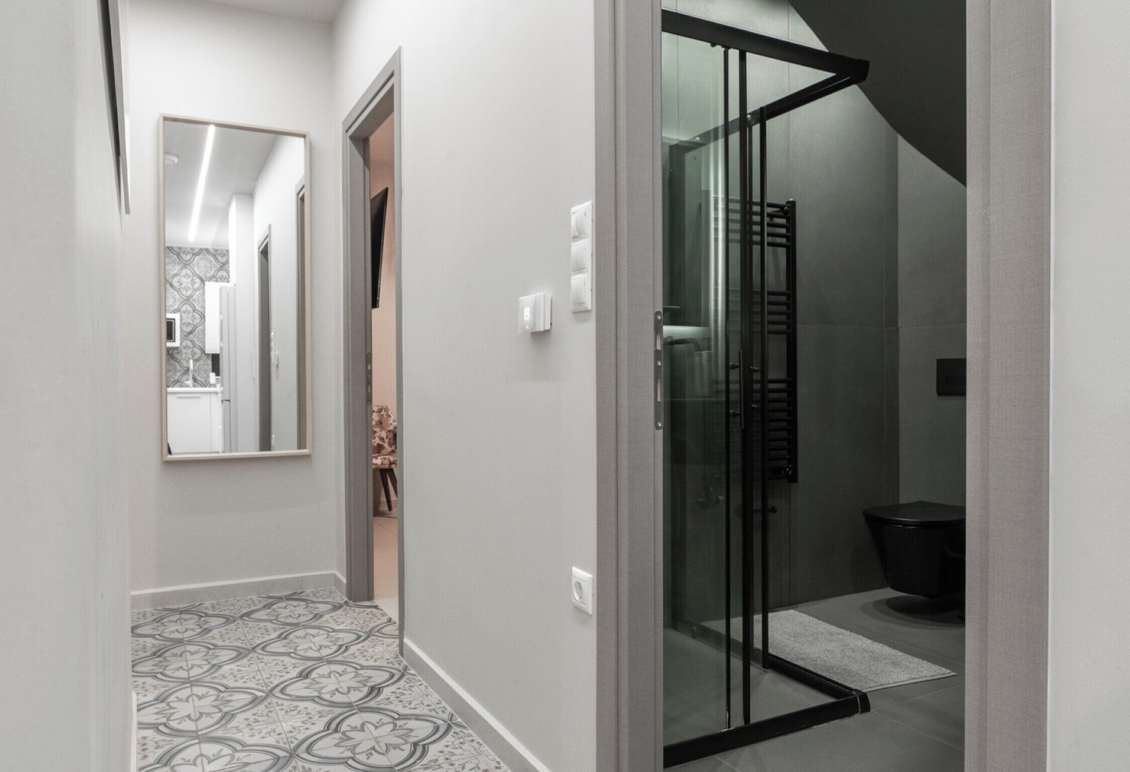 Un pasillo con suelo estampado, un espejo, una puerta de cristal que da a un baño y una pared con azulejos.