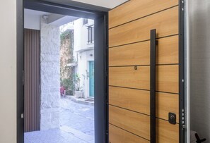 Une porte ouverte donnant sur une cour bordée d’un mur de pierres, d’un mur en panneaux de bois et d’un cadre de porte en métal.