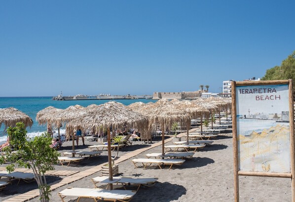A beach with sun loungers and thatched umbrellas, a sign reading 'Ierapetra Beach,' and a view of the sea and distant buildings.