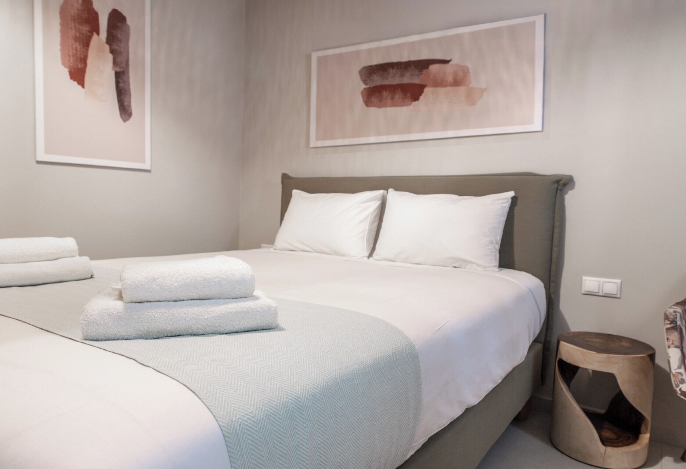 Een opgemaakt bed met witte lakens en kussens, een nachtkastje met een lamp, en kunstwerken aan de muur.