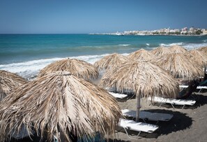 A beach with straw umbrellas and lounge chairs, a clear blue sea, and buildings in the background. - A modern apartment in the center of discreet luxury and comfort 30m from the sea (Ierapetra)