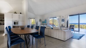 A modern living space with a dining area, sofa, and large windows offering a view of the sea. - Villa Gecko Di Santa Lucia (Zonza)