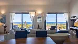 A spacious living room with a large wooden table, striped sofas, and a view of the ocean. - Villa Gecko Di Santa Lucia (Zonza)