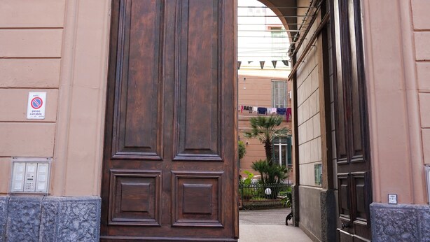 A large wooden door with a metal arch above it, leading to a building with a tiled roof and a small palm tree in the background. - Napoli Incanto (Naples)