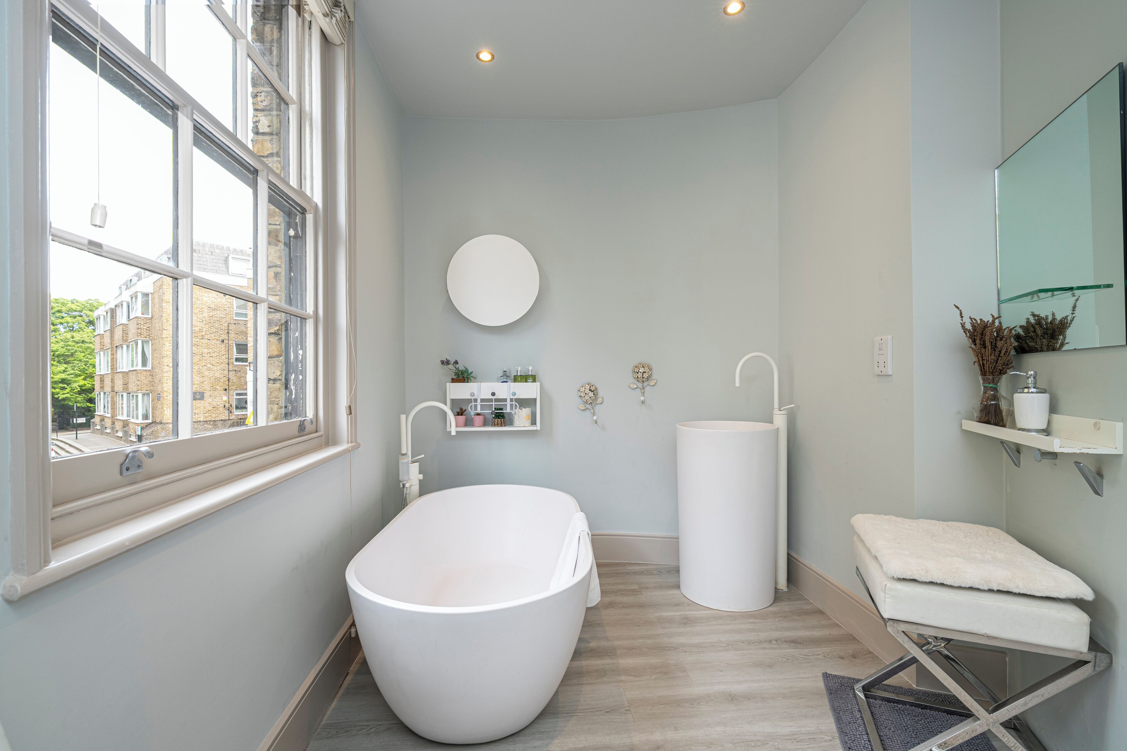 Une salle de bain moderne avec une baignoire sur pieds, un grand miroir et une fenêtre donnant sur les bâtiments environnants.