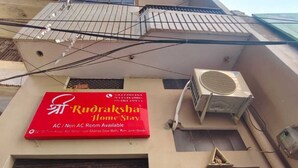 A building with a red signboard advertising AC/Non AC rooms, a large air conditioning unit, and a wooden door with a yellow sign. - Shree Rudraksha Home Stay (Varanasi)