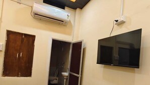 A room with a television mounted on the wall, an air conditioning unit, a wooden door, and a small window. - Shree Rudraksha Home Stay (Varanasi)