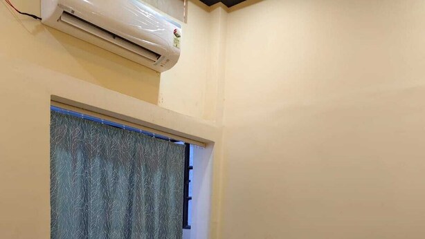 A single bed with a dark headboard, a window with a patterned curtain, and an air conditioning unit mounted on the wall. - Shree Rudraksha Home Stay (Varanasi)