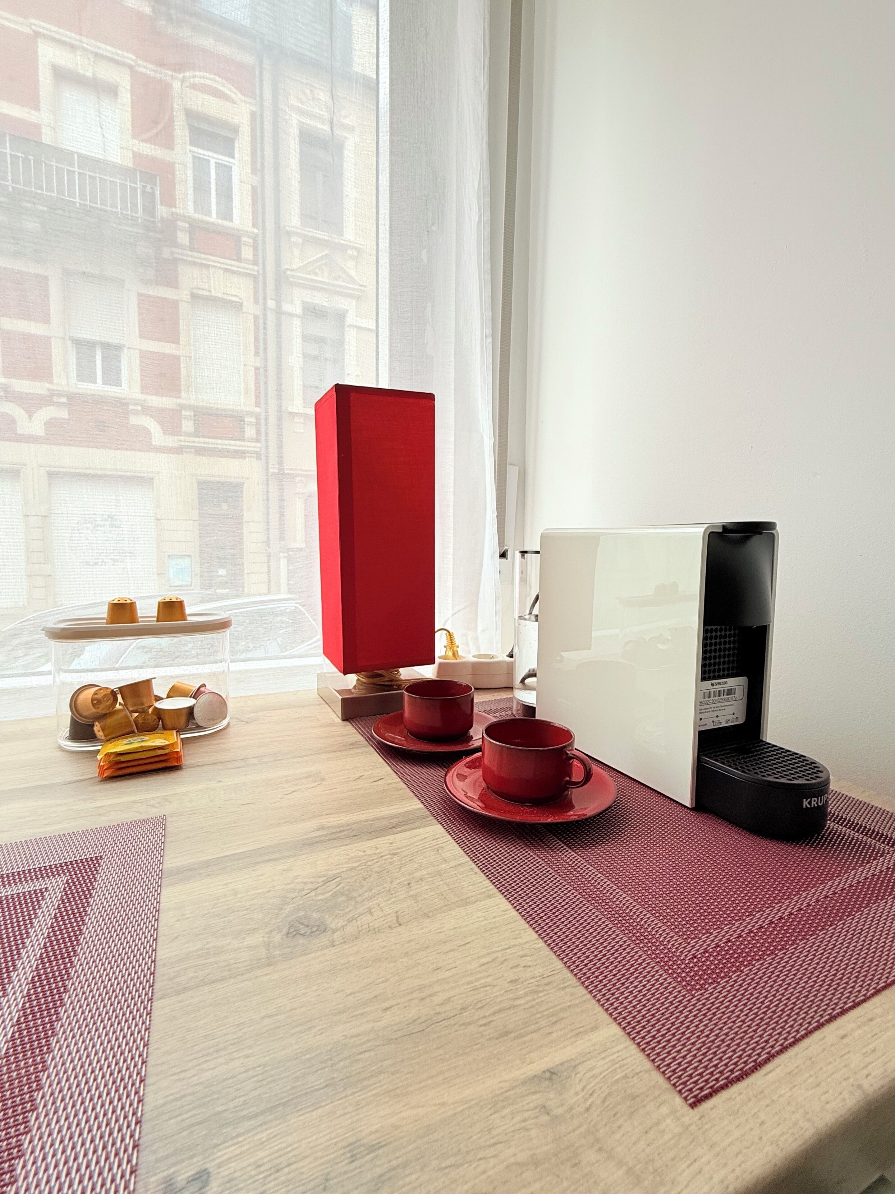 A room with a wooden table, a red box, a coffee maker, and a view of buildings through a window.