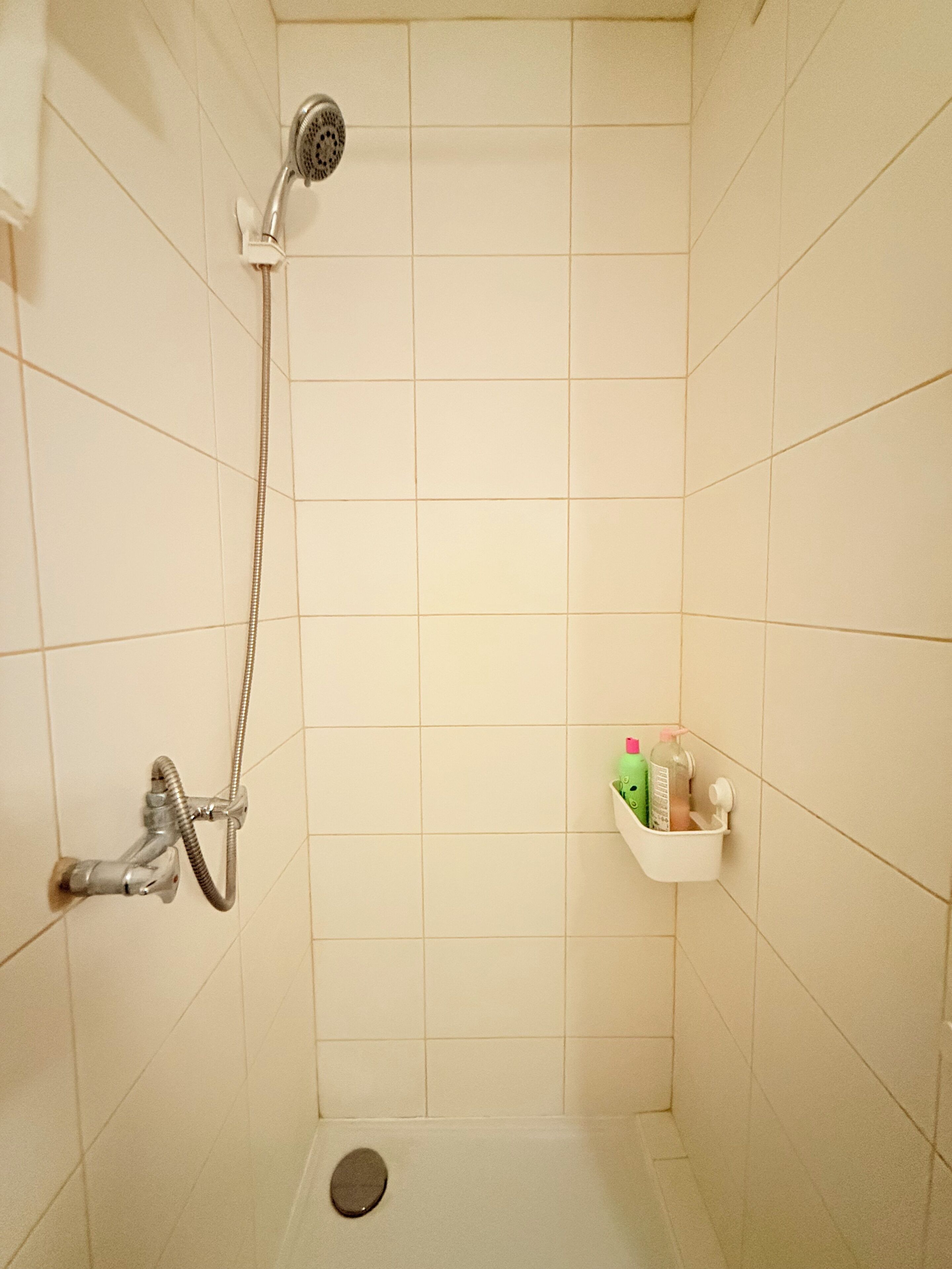 Una ducha con cabezal de ducha de mano, grifo empotrado en la pared y un pequeño estante con artículos de aseo.