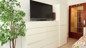 A room with a flat-screen TV on a white cabinet, a potted plant, a bed with a floral bedspread, and a wooden door. - 2 bedrooms - baby crib & furnished  (Luxembourg City)