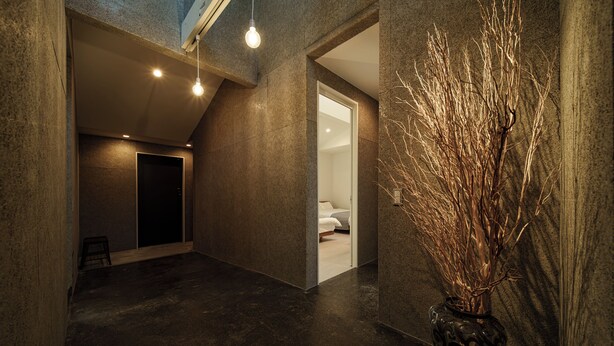 A modern interior with a dark floor, a vase with branches, and a doorway leading to a room with a bed. - Furugo BASE (Oita)