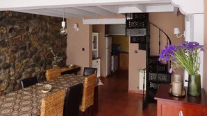 Un comedor con pared de piedra, una mesa de comedor con sillas de mimbre, una escalera y una cocina al fondo.