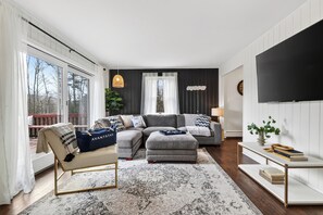 Ein modernes Wohnzimmer mit einer Eckcouch, einem kleinen Beistelltisch mit Büchern, einem großen Fernseher und Blick ins Freie durch eine Schiebetür.