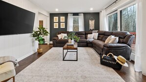 A living room with a sectional sofa, a coffee table, a TV, and a large window. - 6BR + Hot Tub + Firepit + Playroom | Mins to Hunter Mountain + Catskills Hikes (Tannersville)