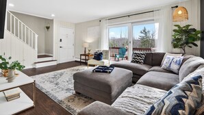 A living room with a sofa, ottoman, and a staircase. - 6BR + Hot Tub + Firepit + Playroom | Mins to Hunter Mountain + Catskills Hikes (Tannersville)
