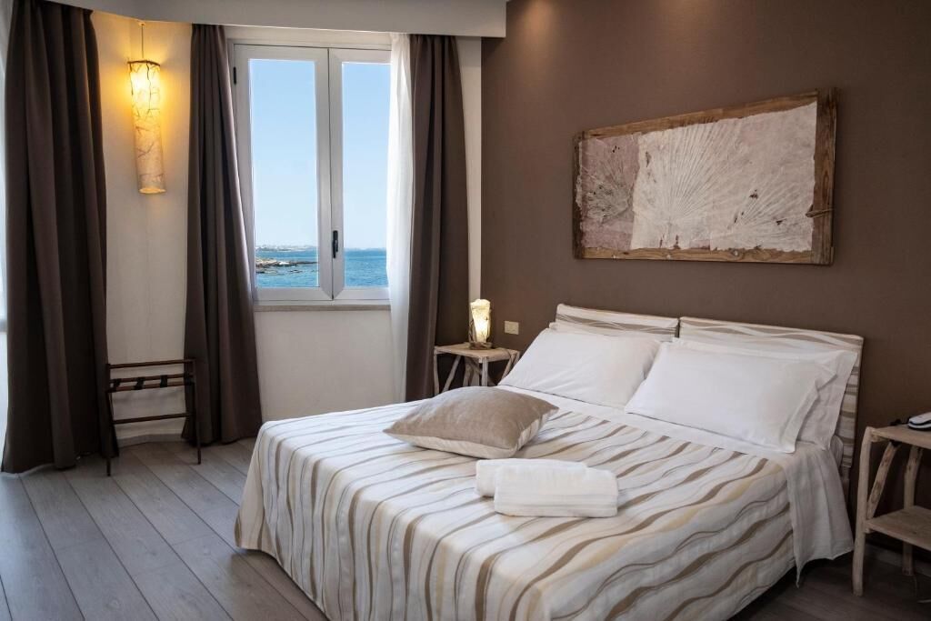 Ein Hotelzimmer mit Bett, Nachttisch, Lampe, einem Fenster mit Vorhängen und einem Bild an der Wand.