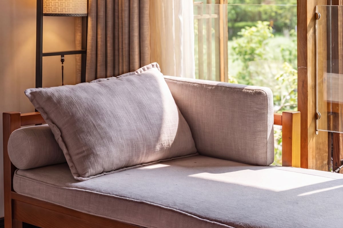 Een kamer met een houten bankje, een wandlamp en uitzicht op groen via een deur.