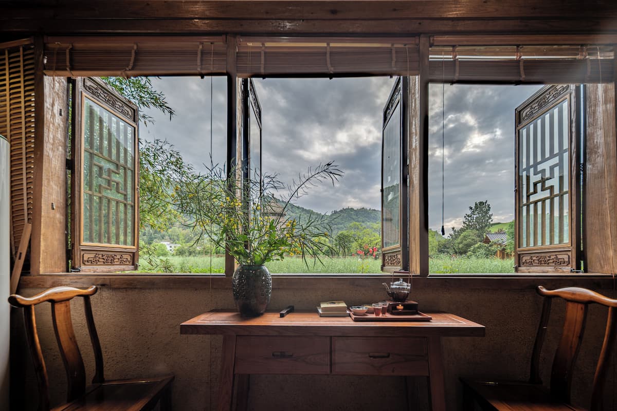 Een kamer met een bureau, twee stoelen, een vaas met bloemen en uitzicht op groen en bergen door grote ramen.