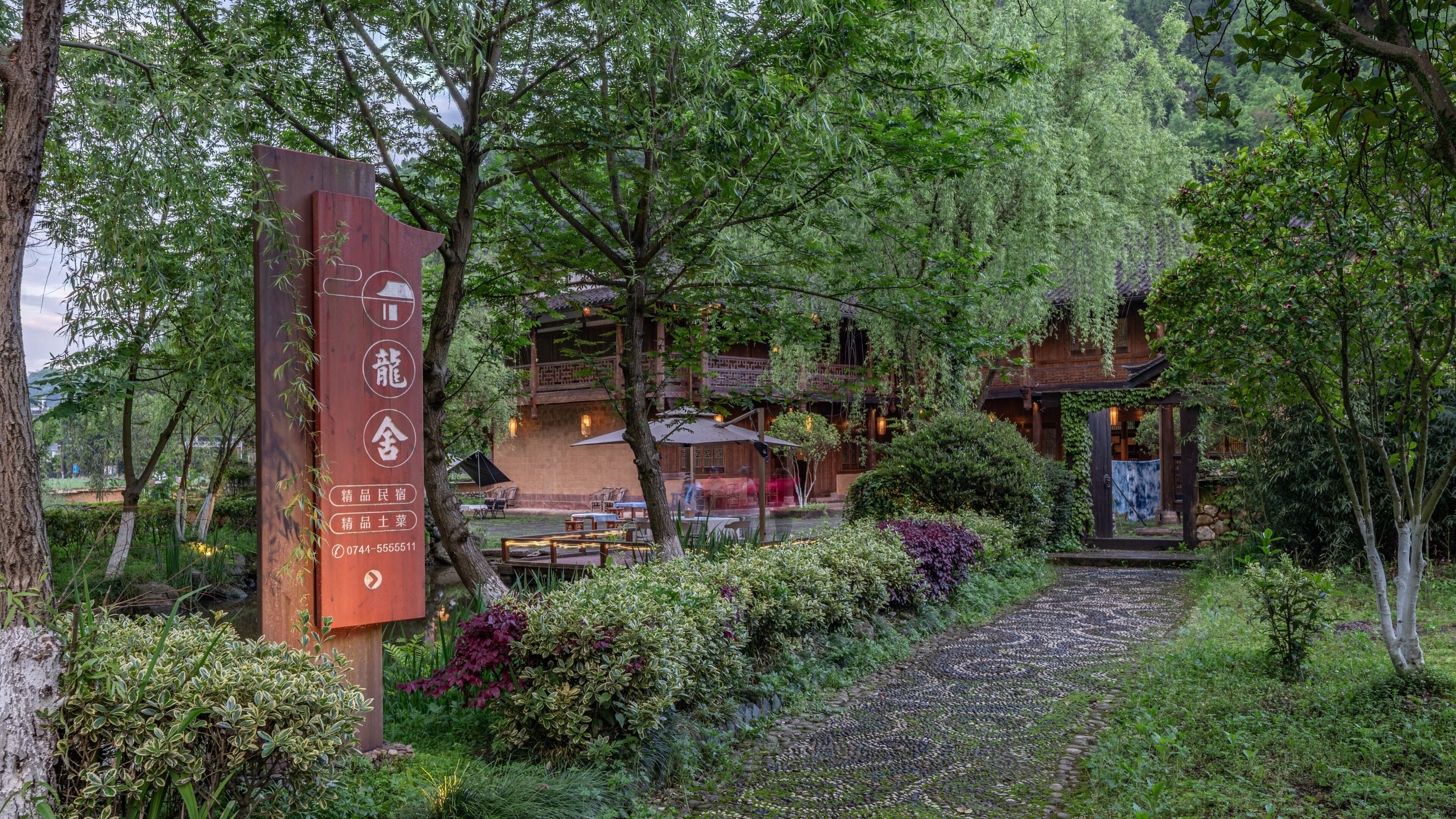 A traditional-style building with a wooden exterior, surrounded by lush greenery and a stone pathway leading to the entrance.