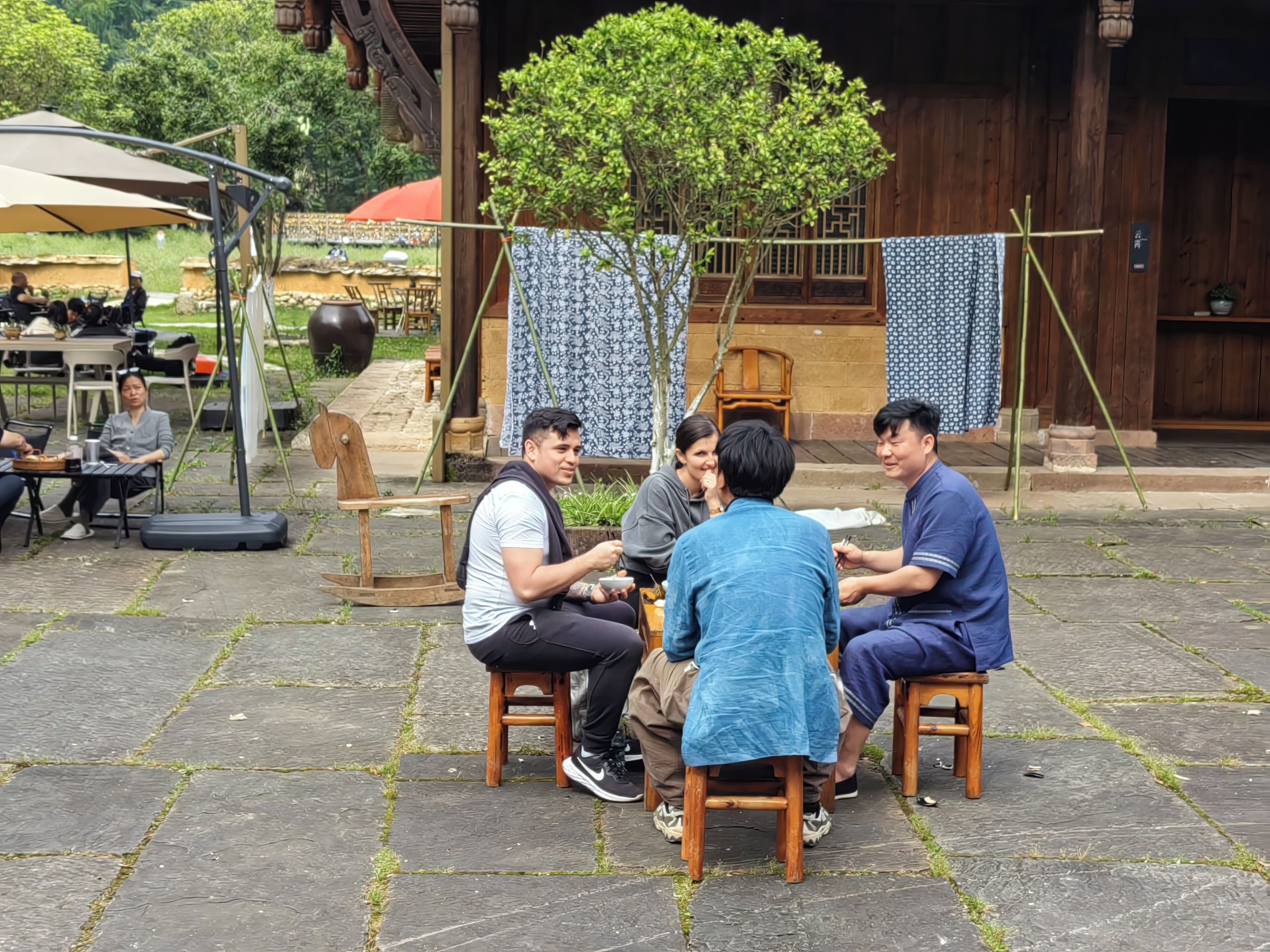A group of people seated at a table outdoors, with a wooden structure and a tree in the background.