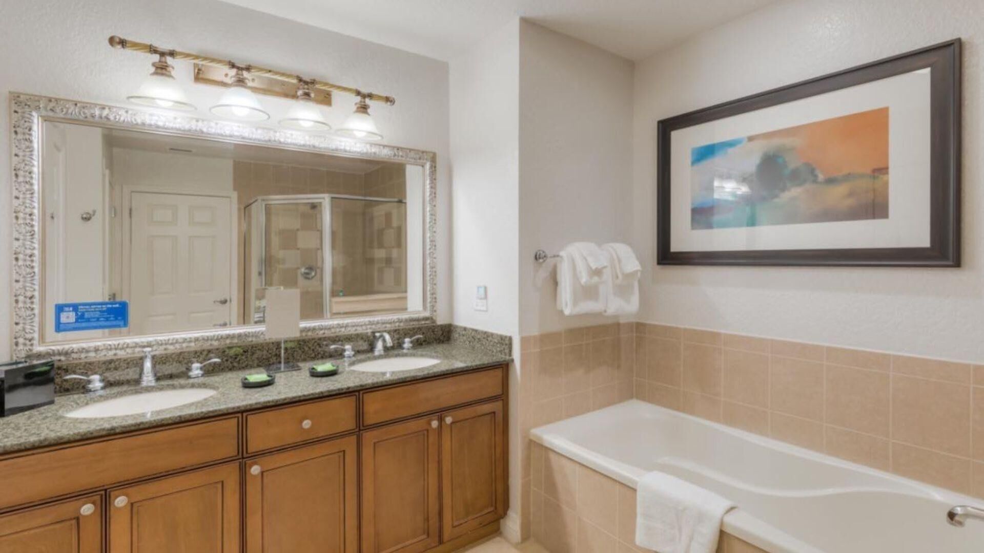 A bathroom with a double sink, a large mirror, a framed picture on the wall, and a bathtub.