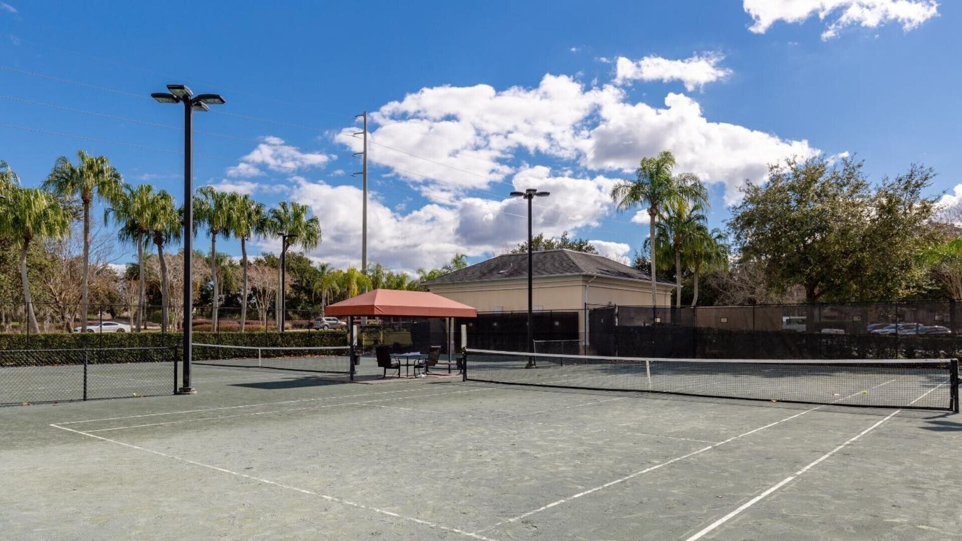 Un court de tennis en plein air doté d’une zone de sièges couverte, entouré de palmiers et avec un bâtiment en arrière-plan.