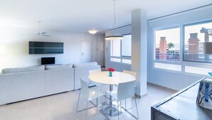 En moderne stue med sofa, spisebord med stole, fjernsyn ophængt på væggen og et stort vindue med udsigt til bygninger.