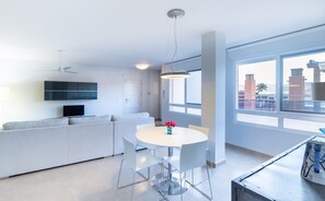 A modern living room with a sofa, a dining table with chairs, a TV mounted on the wall, and a large window with a view of buildings. - Stunning Luxury Penthouse with Sea Views, Wi-Fi and Air Conditioning (La Azohía)