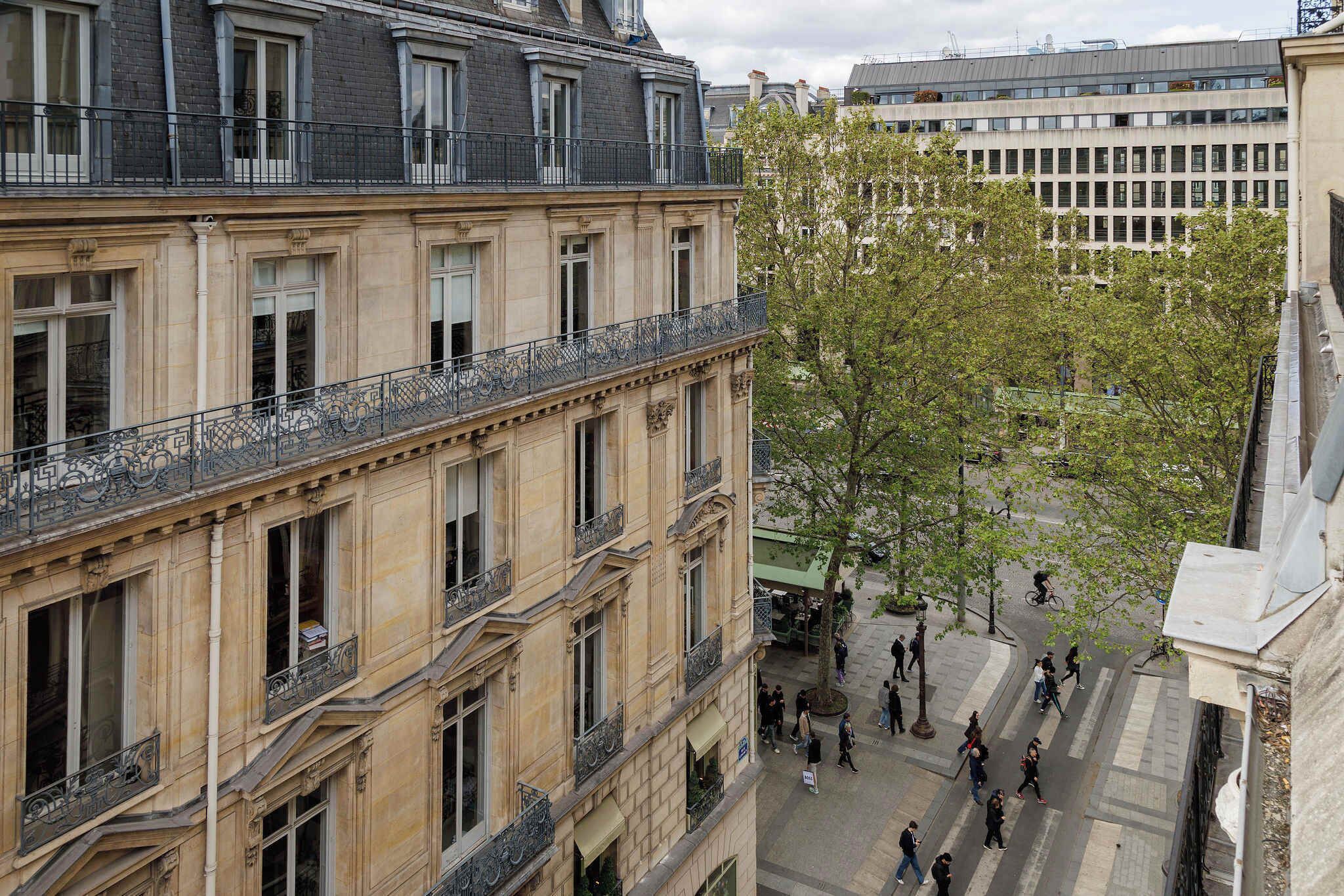A multi-story building with balconies, a pedestrian area with trees, and a busy street below.