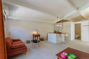 Een moderne woonruimte met een bank, een kleine tafel met lamp, een keukenhoek en een eettafel met stoelen.